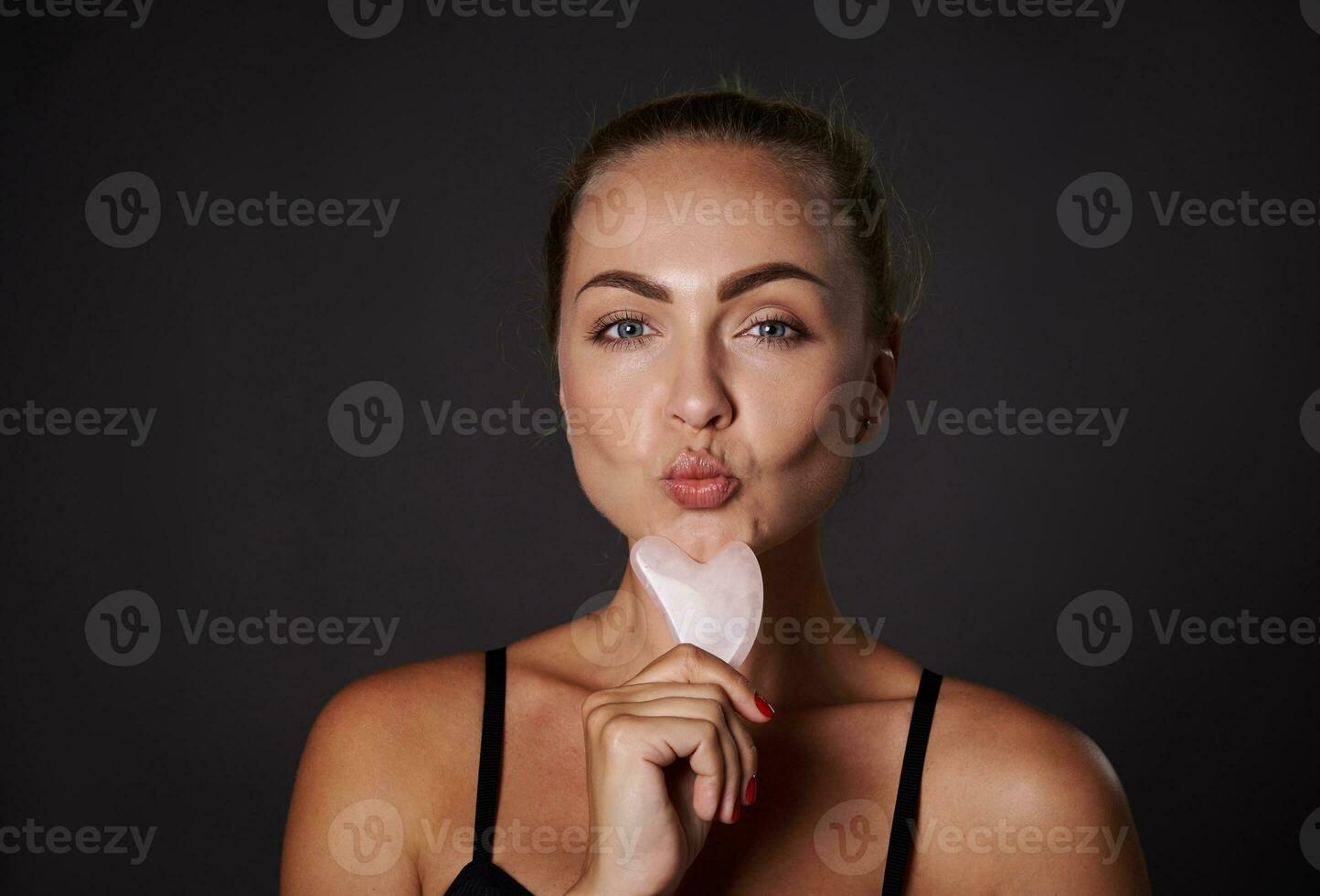 Beautiful Caucasian woman with healthy fresh smooth tanned skin posing with a gua-sha, ayurvedic jade stone massager for facial lifting and lymphatic drainage massage, isolated over black background photo