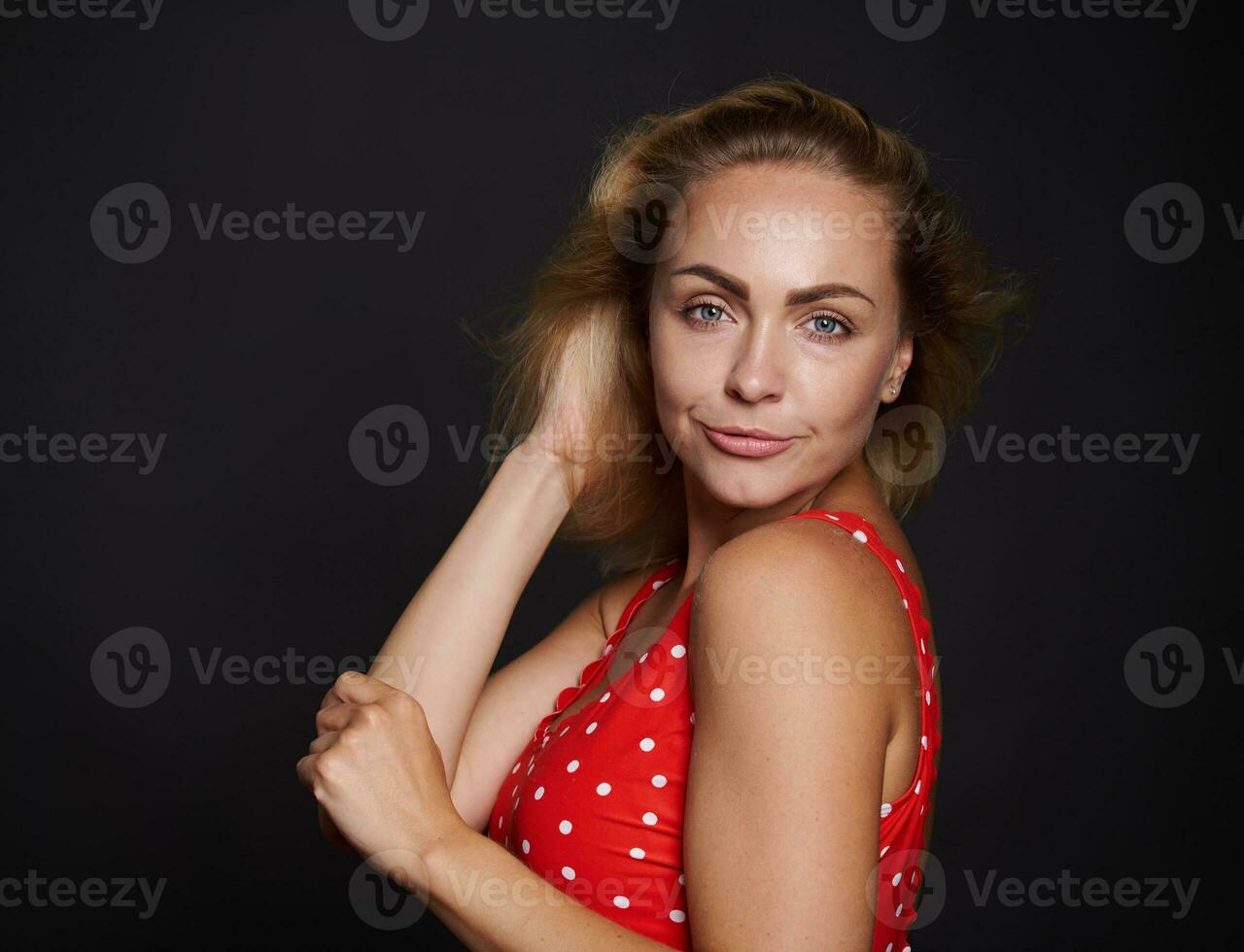 Beautiful middle aged blonde sporty European woman in red swimsuit with white polka dots confidently looking at camera isolated over black background with copy ad space photo