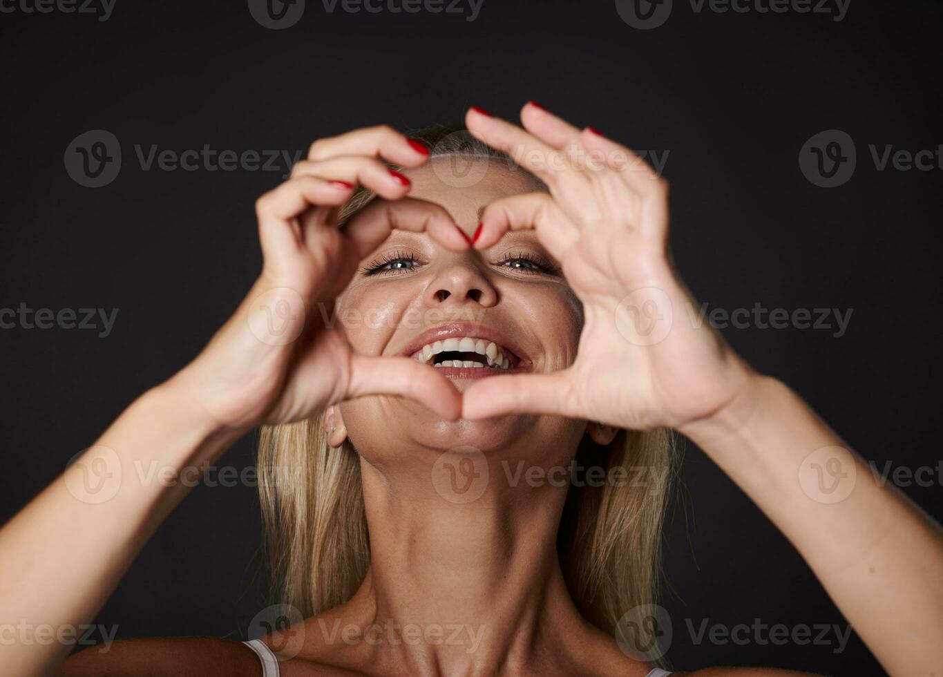 Close-up portrait of a cheerful good-looking blonde Caucasian woman smiles with beautiful healthy toothy smile looking at camera through a finger frame in heart shape, isolated over black background photo