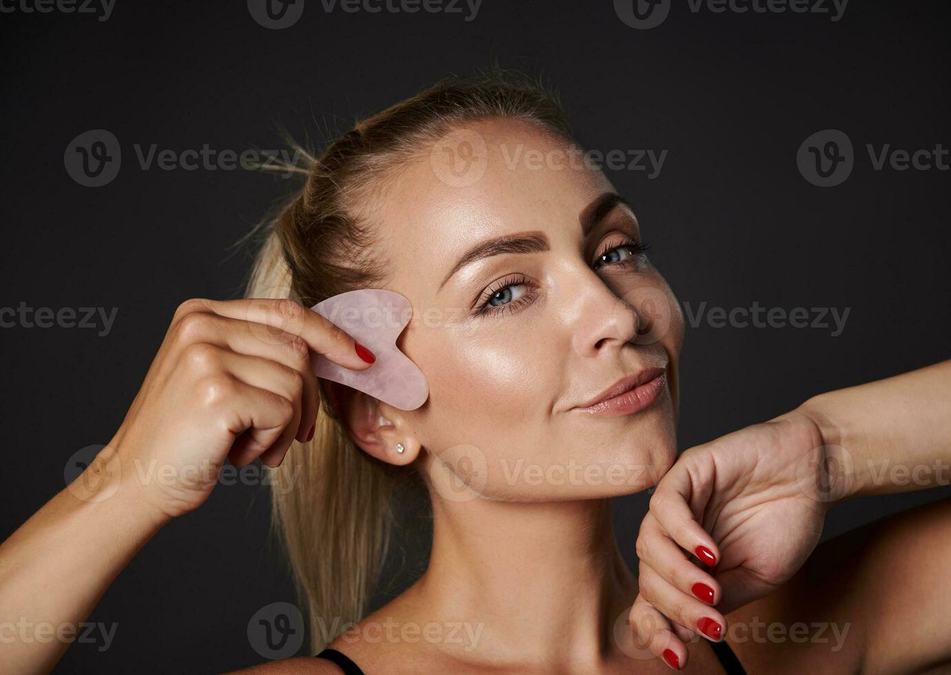 Close-up beautiful woman doing facial lifting smoothing lymphatic drainage massage on her face, using a gua-sha jade stone roller massager, caring of her skin health and youth photo