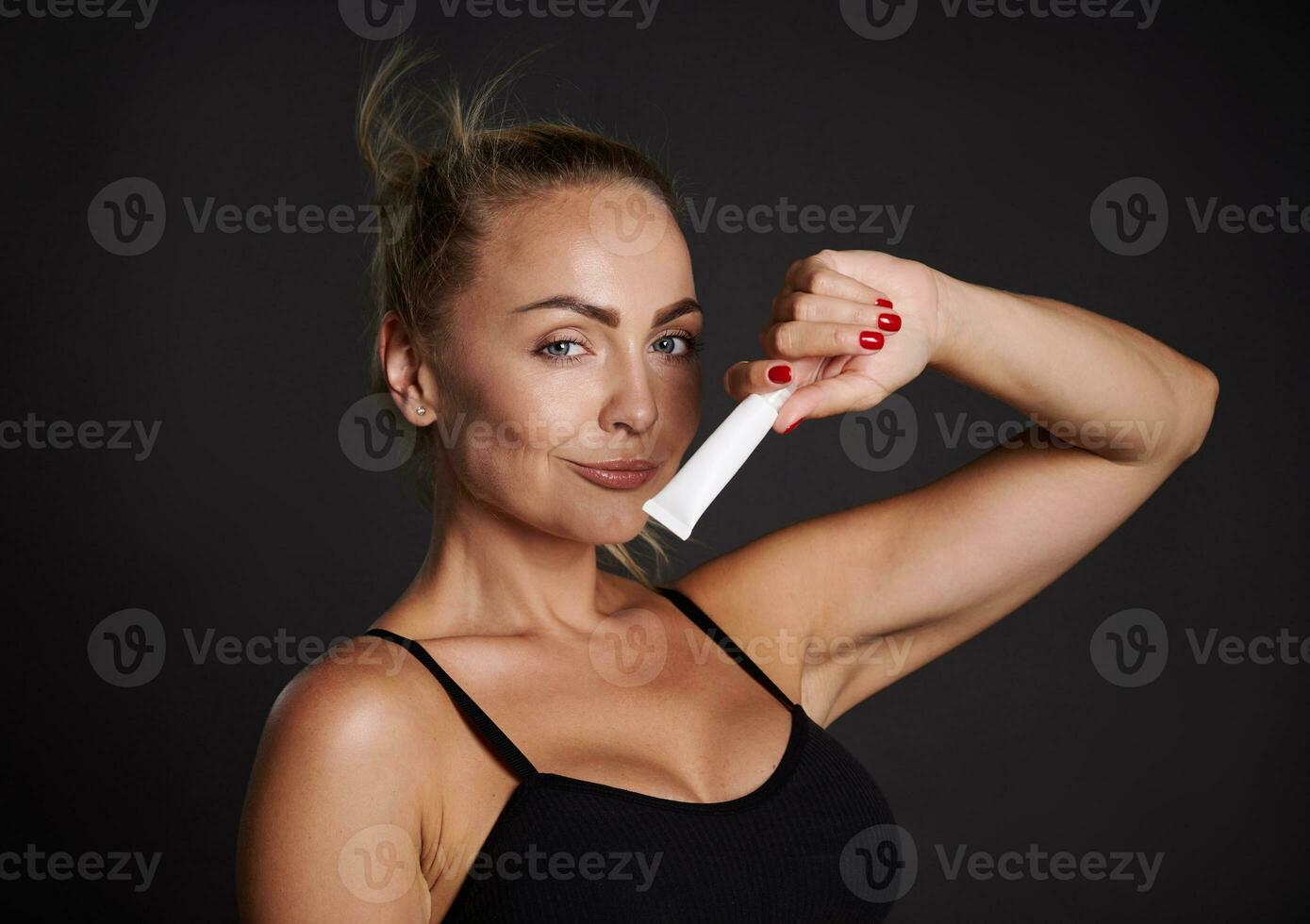 Beautiful Caucasian woman with tanned skin holding a tube of anti wrinkles smoothing anti puffiness under eyes cream, smiles looking at camera, isolated over black background with copy ad space photo