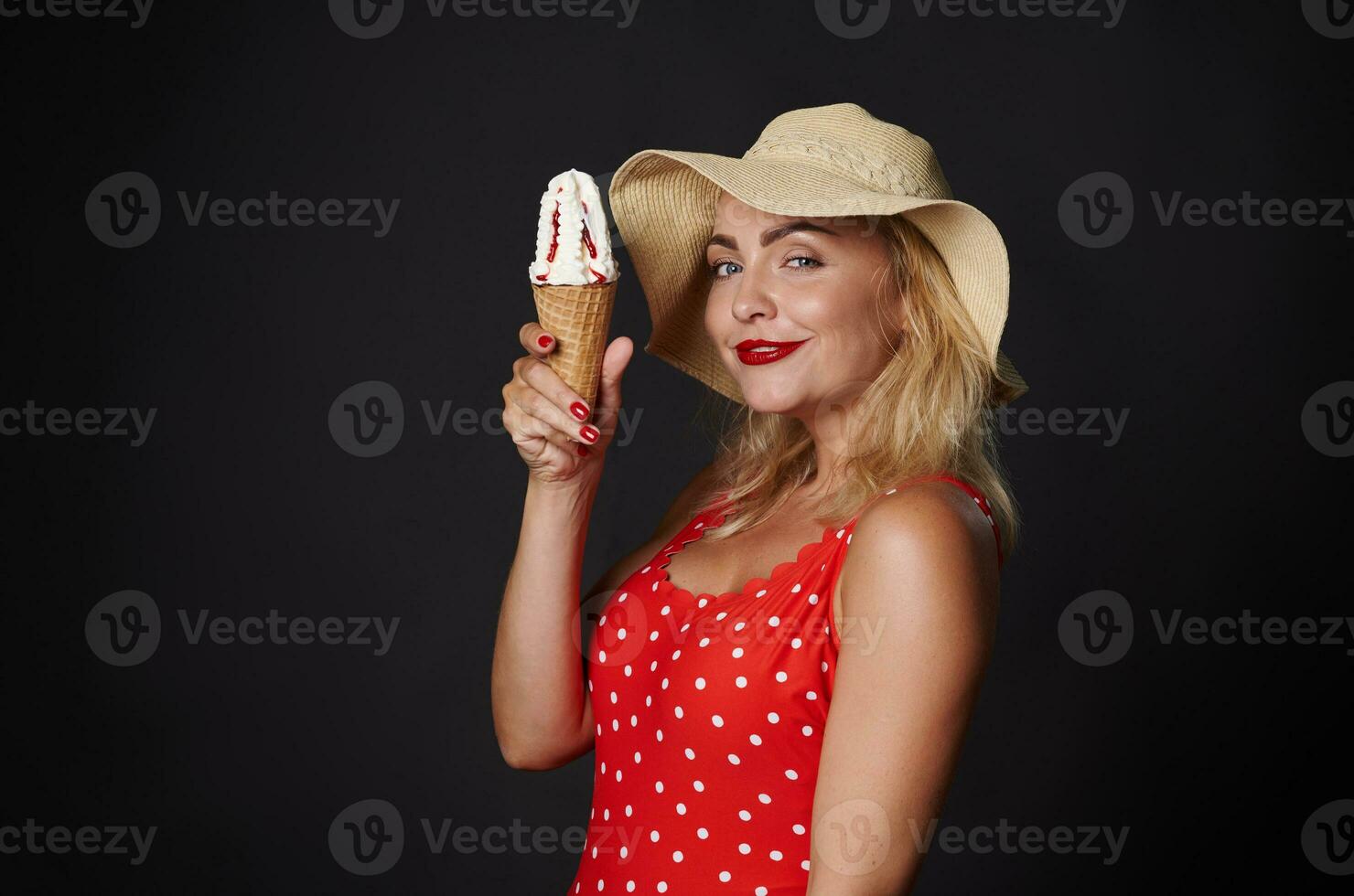 Attractive blonde woman in red swimsuit with white polka dots, holding delicious ice cream cone , smiling beautiful toothy smile, posing on black background copy space photo