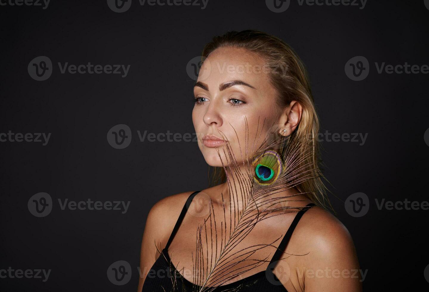 Beautiful middle aged blonde woman with healthy fresh clean skin holding a peacock feather, isolated over black background with copy ad space photo