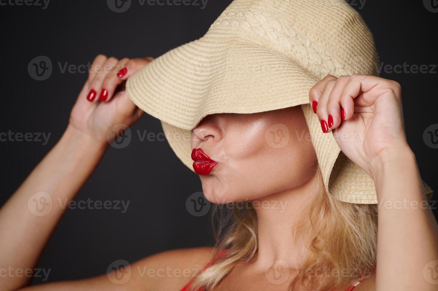 Close-up beautiful Caucasian woman covering her eyes with a straw summer hat and sending kiss, isolated over black background with copy space. Headshot portrait of a pretty lady with red lipstick photo