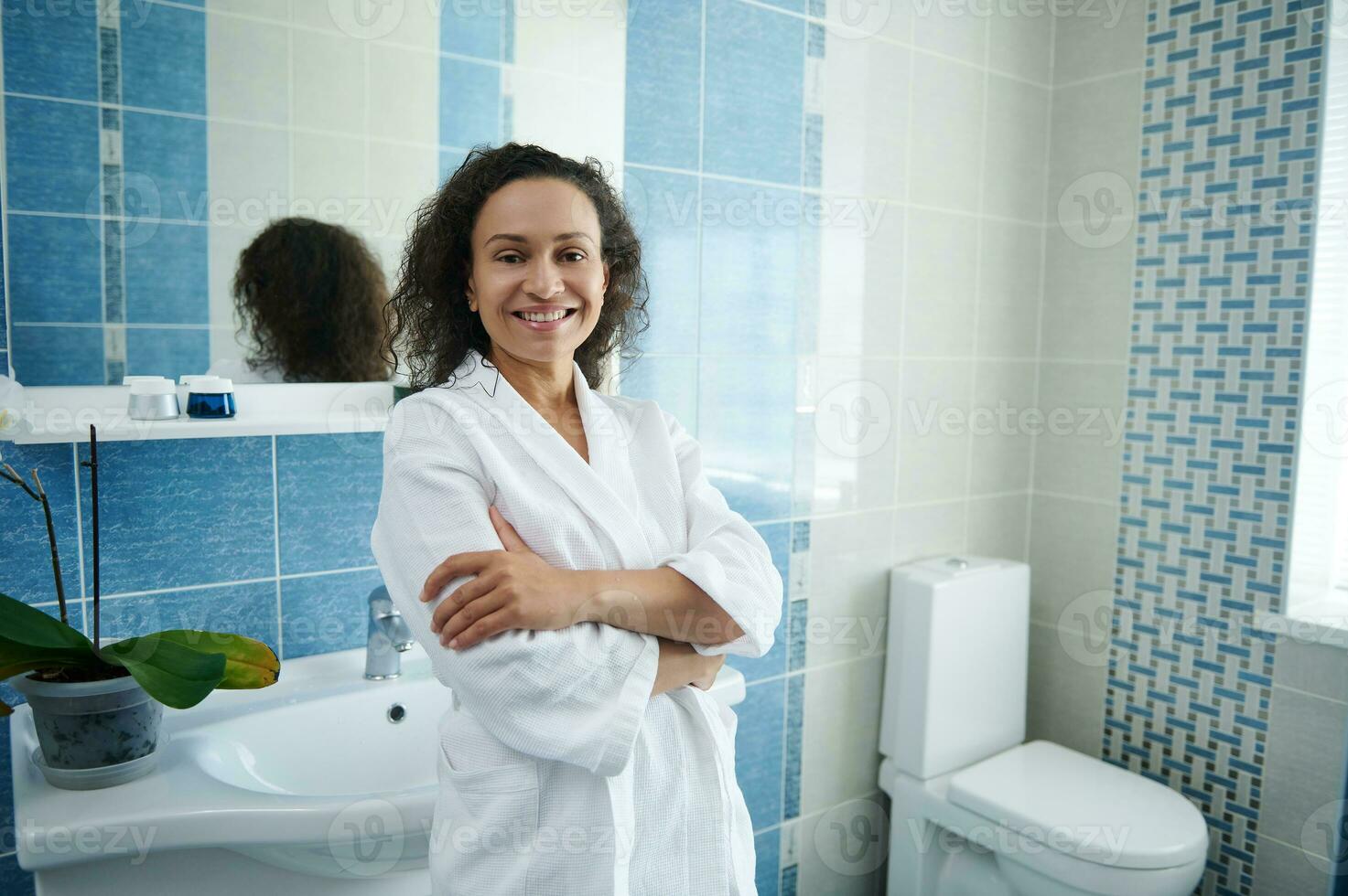 Mature African American woman in white waffle bathrobe leaning on sink in bathroom and smiling at camera with crossed arms photo
