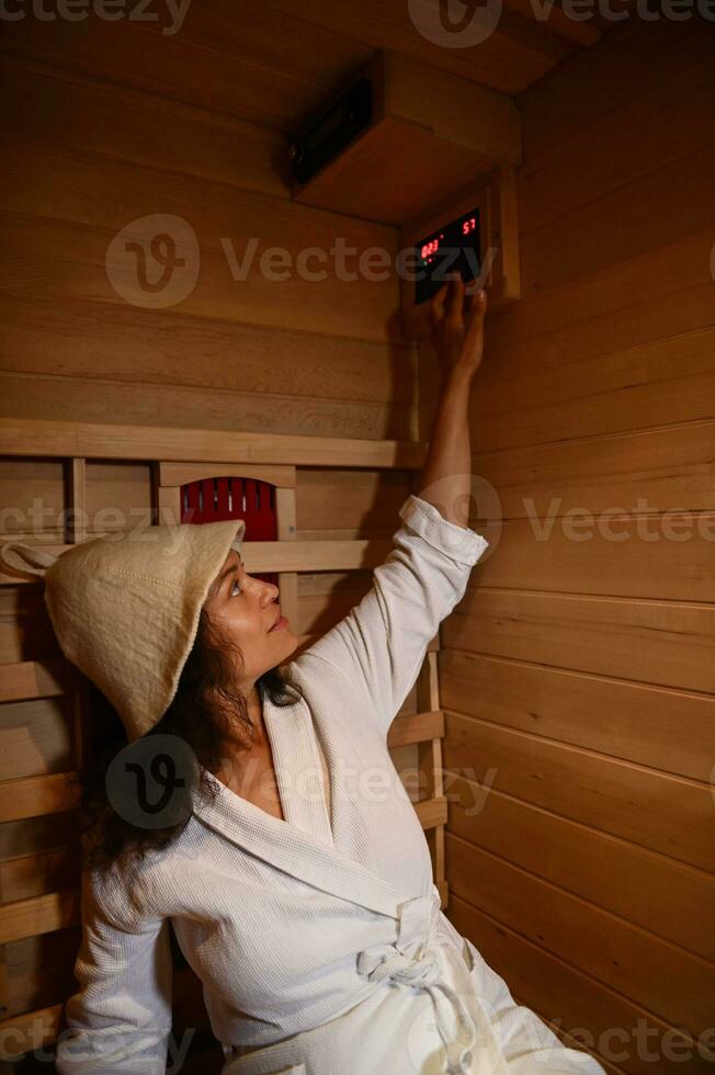 Happy woman wearing a bathrobe and a sauna hat regulates the temperature in the infrared sauna photo