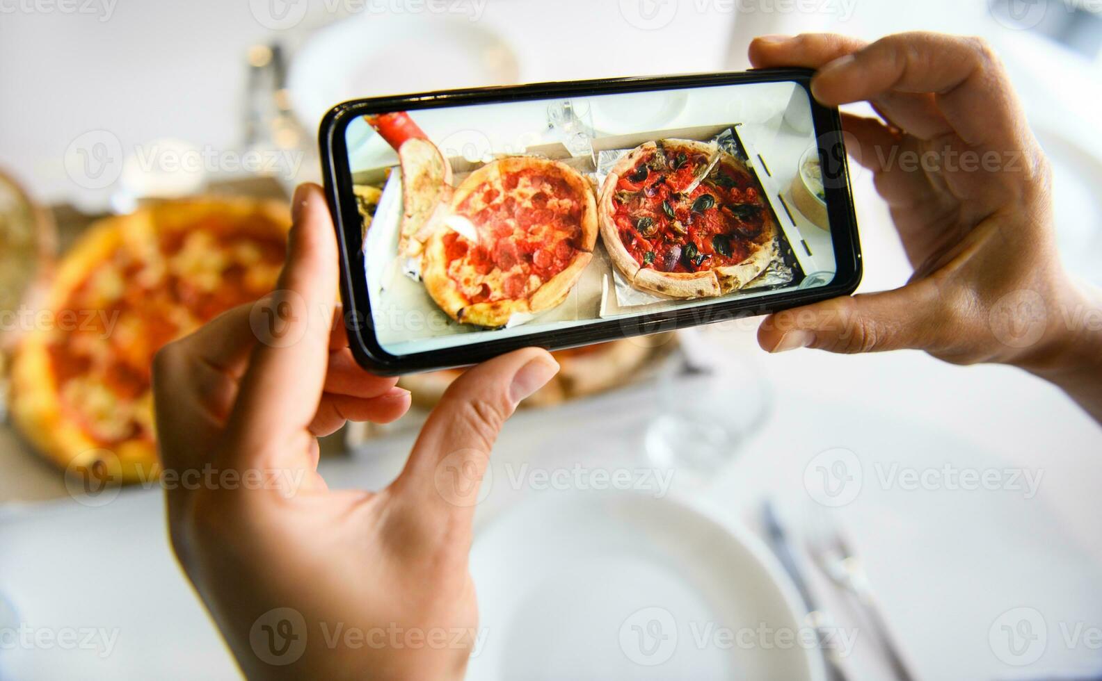 Hands holding smartphone and taking image of delicious freshly baked pizza served on a table with white tablecloth. Mobile phone in live mode regime photo