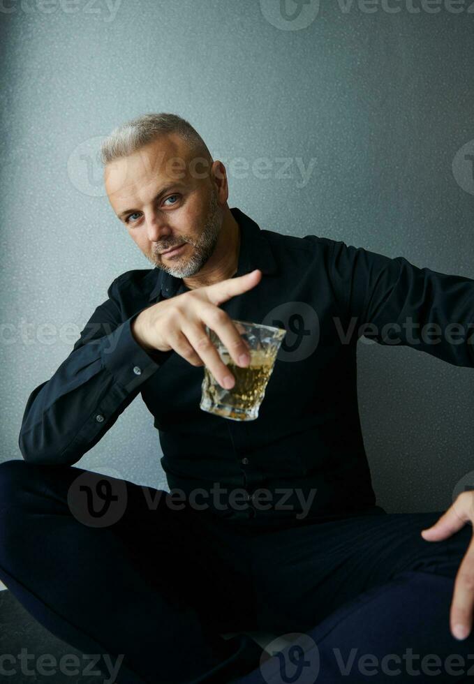 Handsome attractive middle aged Caucasian man in black casual suit, looking confidently at camera posing with a glass with alcoholic drink against gray wall background photo