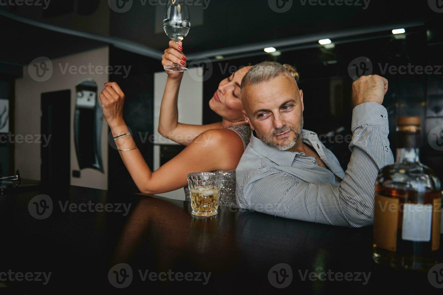 atractivo bronceado europeo mujer y hermoso caucásico medio Envejecido hombre Bebiendo alcohol y teniendo divertido juntos mientras celebrando un evento a hogar bar. coquetear, seducción, Pareja relaciones concepto foto