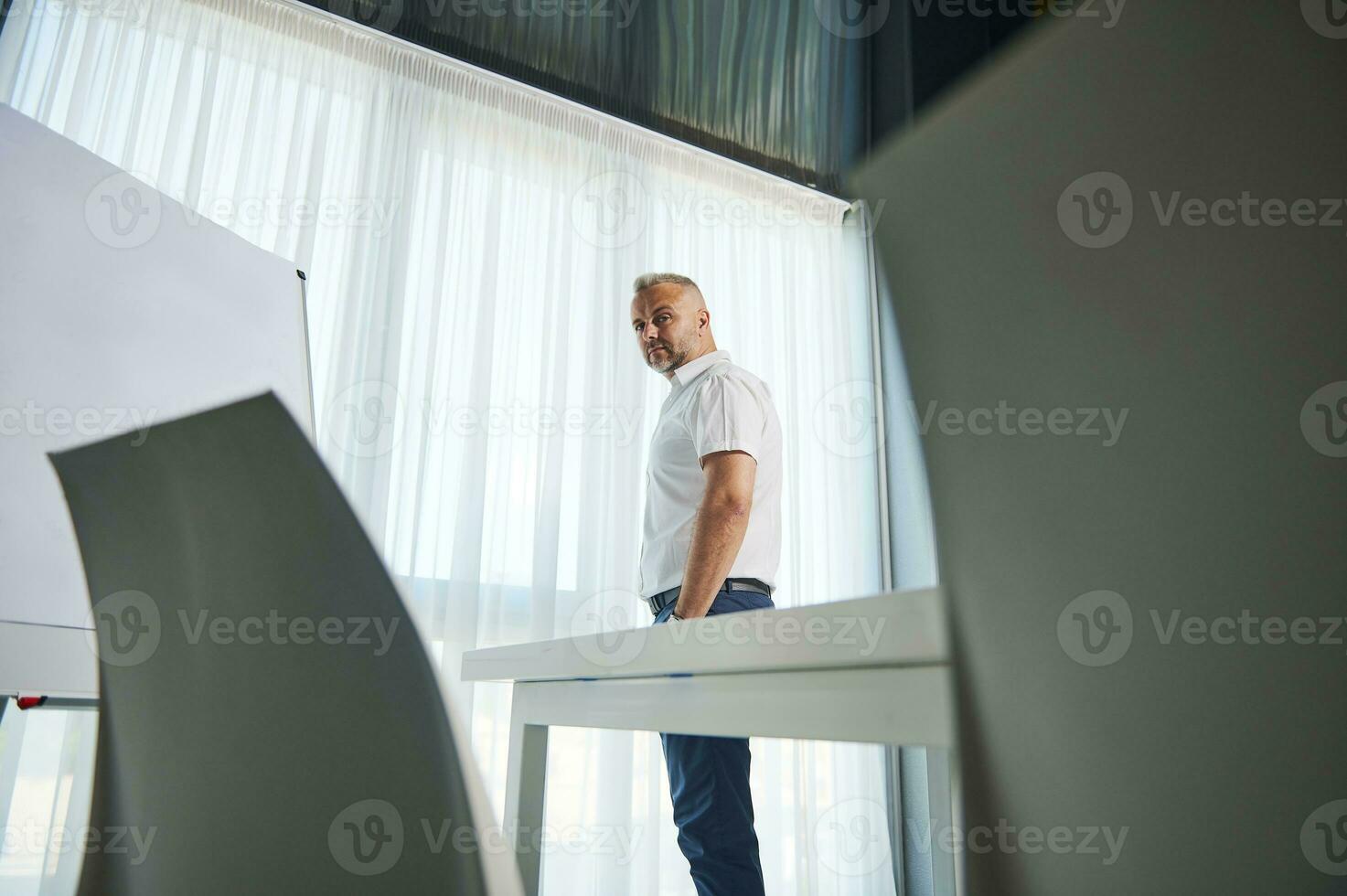 Attractive middle aged entrepreneur, successful sales manager, businessman standing near flip chart with copy space for advertisement text, and confidently looking at camera photo