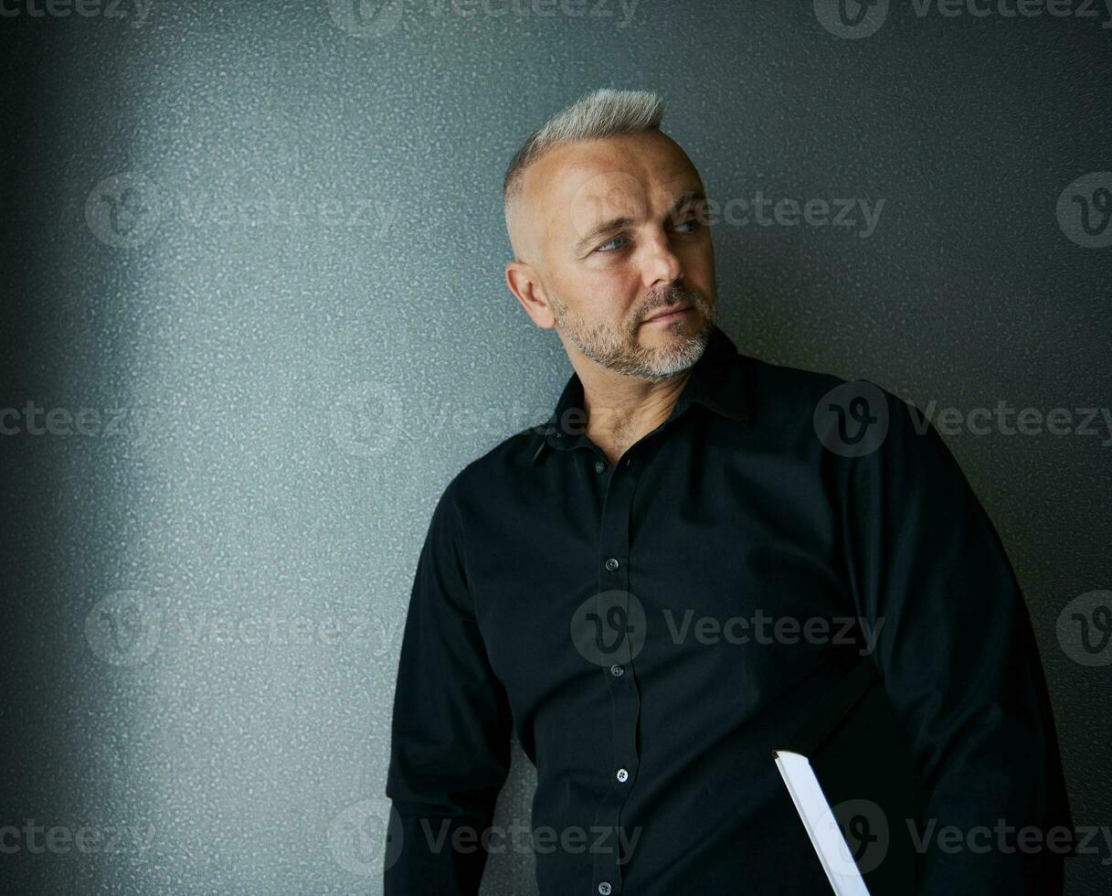 Handsome stylish middle aged Caucasian man, successful businessman, entrepreneur looking aside posing against gray wall background with a book. Copy space photo