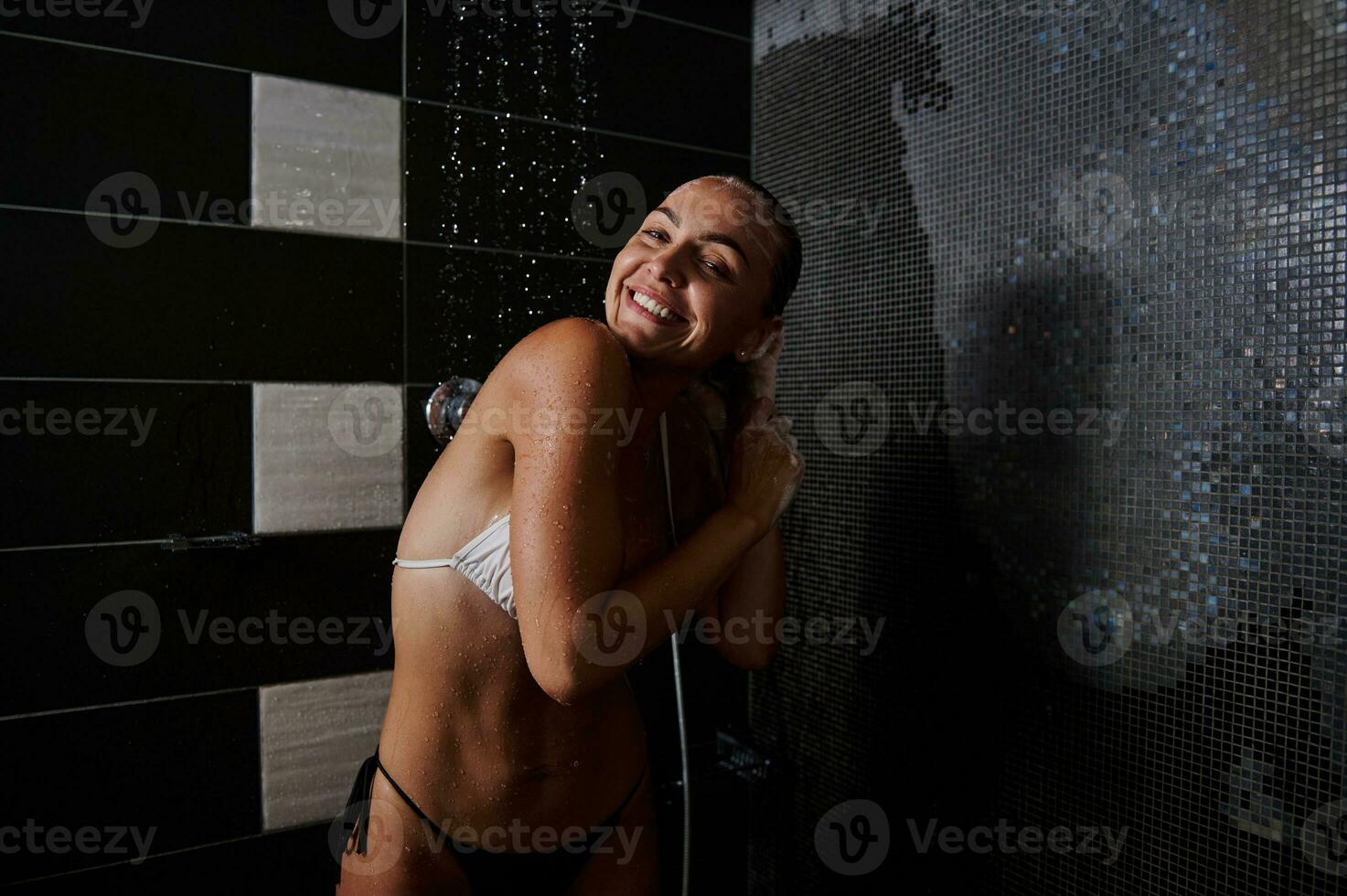 alegre rubia joven mujer lavados cabeza en pie debajo un ducha a hogar baño y lindo sonriente con dientes sonrisa mirando a cámara. pureza, limpieza y cuerpo cuidado concepto foto