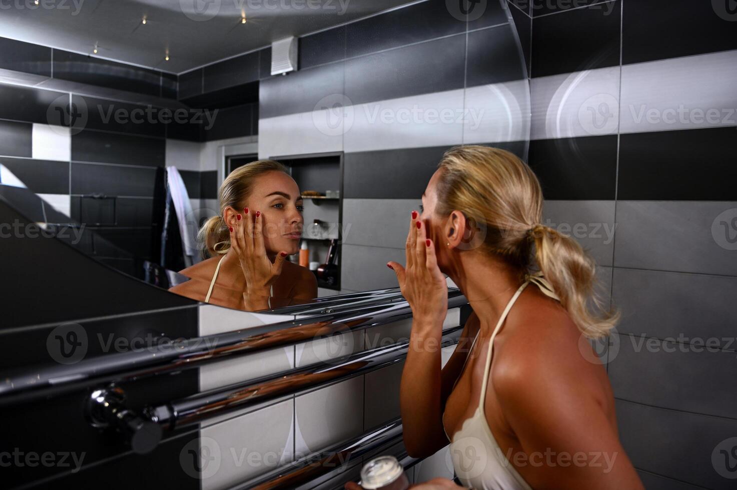 Charming young Caucasian blonde woman in swimsuit applying moisturizer, performing smoothing rejuvenating facial massage looking at her reflection in bathroom mirror photo