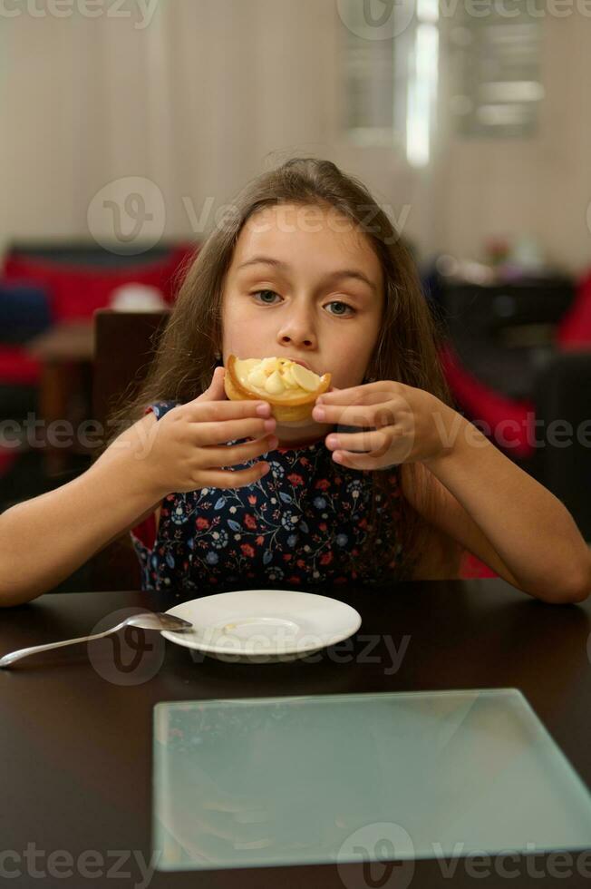 auténtico linda pequeño niño niña sentado a mesa y disfrutando comiendo Fresco dulce azucarado postre - un tartaleta con limón foto