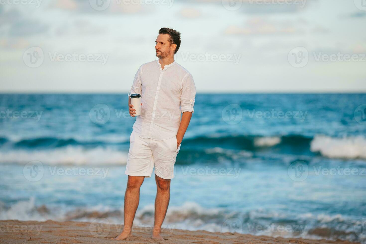 Young man in white with coffee on the beach walking and enjoy evening time photo