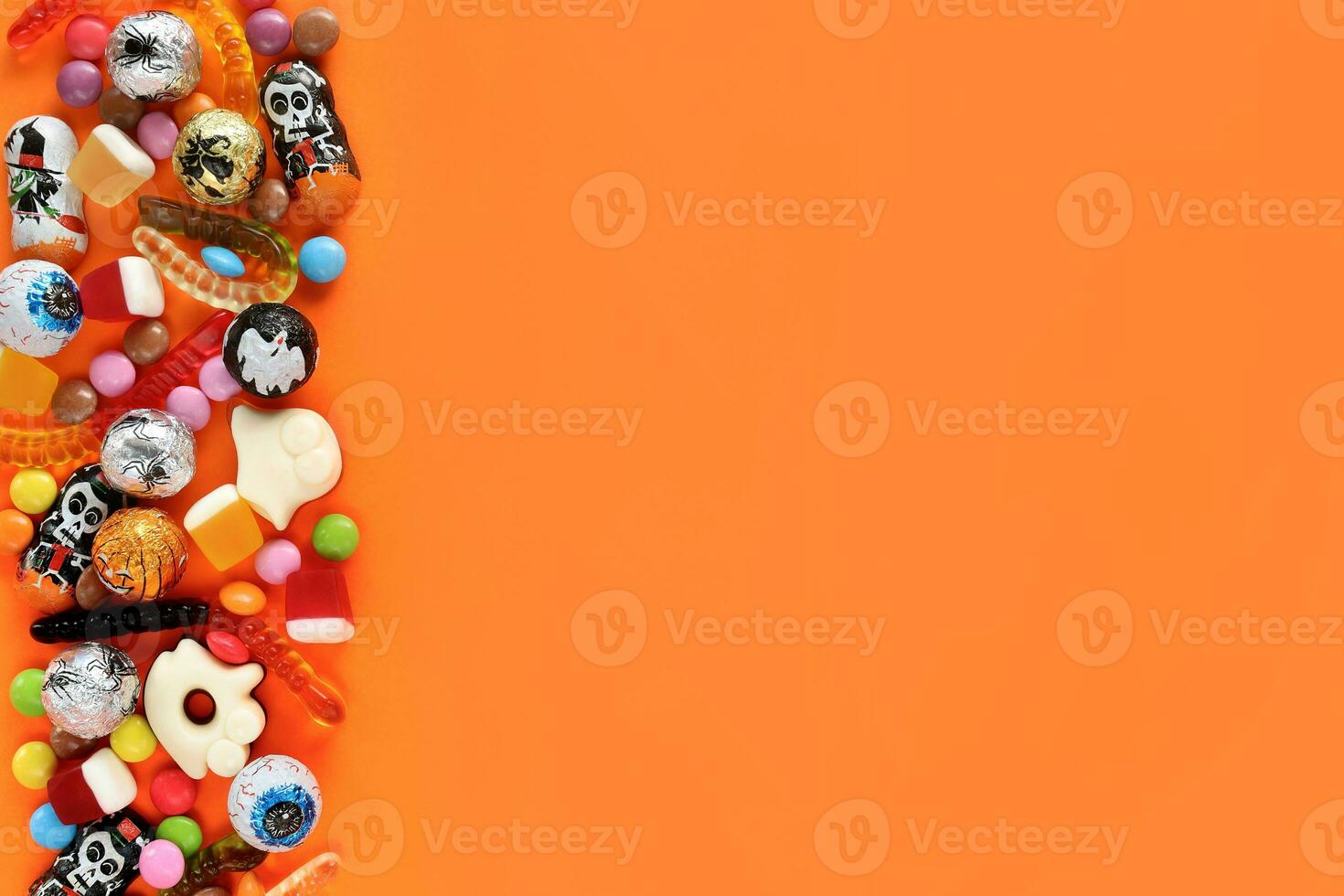 Halloween orange background with copy space on right, assorted sweets on left - traditional eyeballs chocolates, jelly worms, gummy ghosts. Happy Halloween holiday sale and trick-or-treat concept. photo