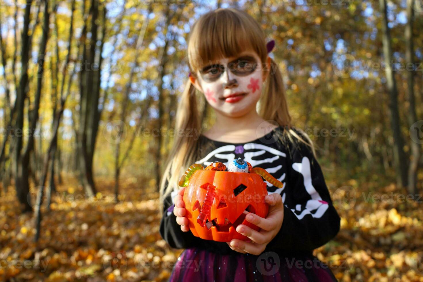 pequeño niña en Víspera de Todos los Santos esqueleto negro vestir con azúcar cráneo constituir, borroso rostro. selectivo atención en manos con naranja calabaza Cubeta lleno de tradicional dulces mascarada disfraz para día de muerto foto