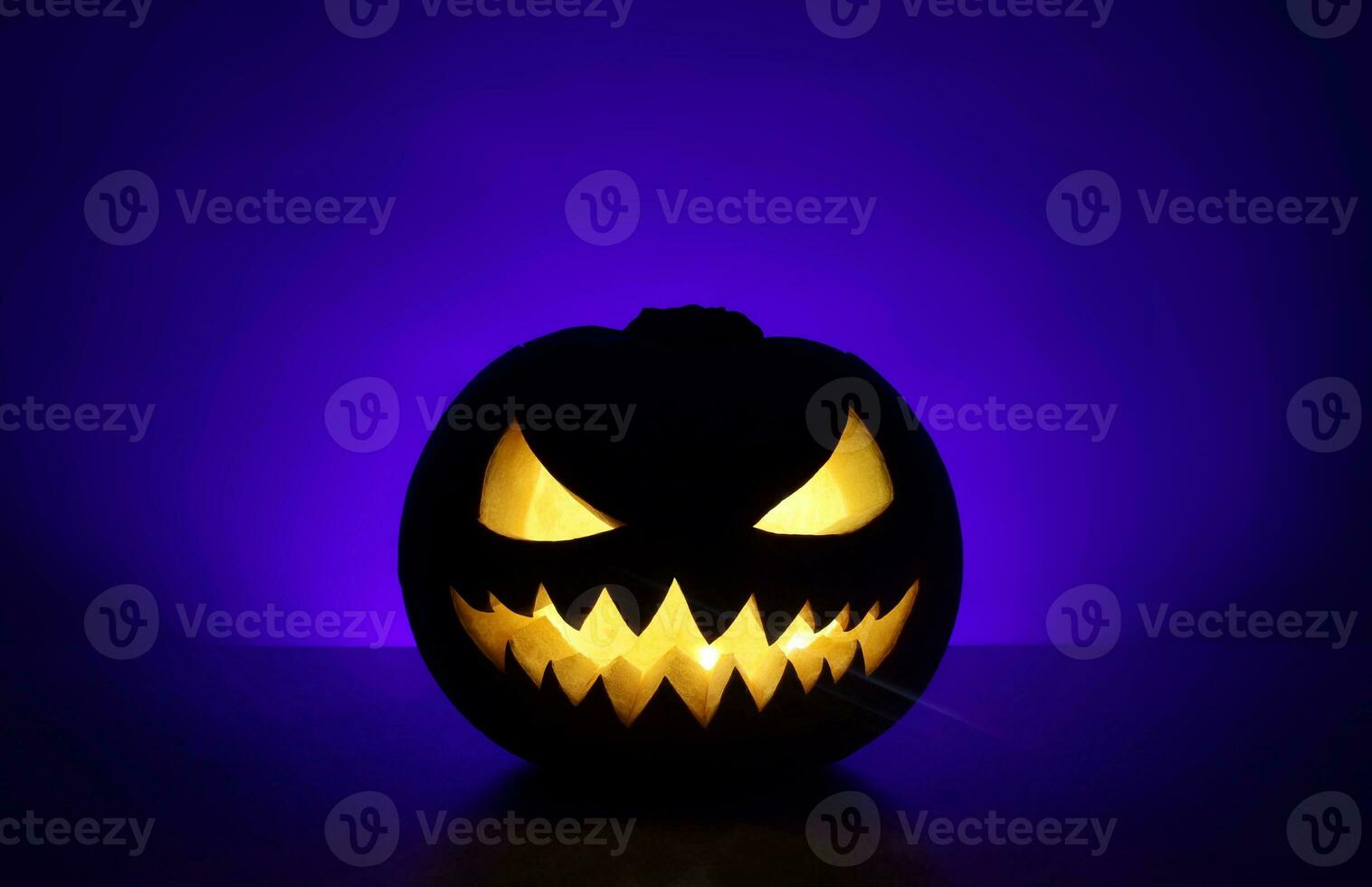 Halloween holiday background with traditional spooky angry jack-o-lantern. Carved glowing pumpkin stands on surface in dark night. Back wall with place for text is highlighted blue with shadows. photo