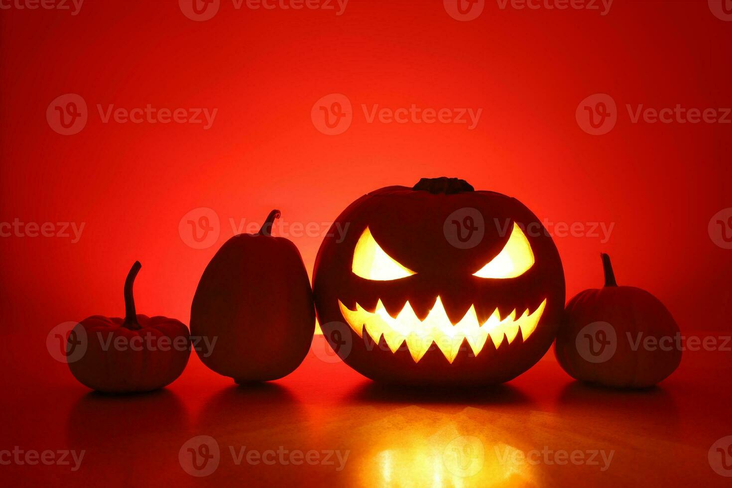 Halloween spooky angry jack-o-lantern and three little pumpkins silhouettes. Carved glowing pumpkin stands on surface in dark night. Back wall with copy space is highlighted red with shadows. photo