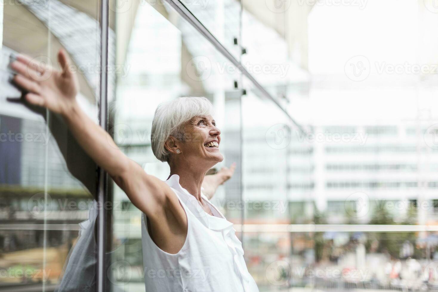 Happy senior woman leaning against glass facade with outstretched arms photo