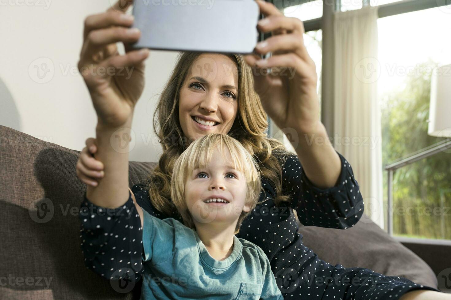 sonriente madre con hijo tomando un selfie en sofá a hogar foto