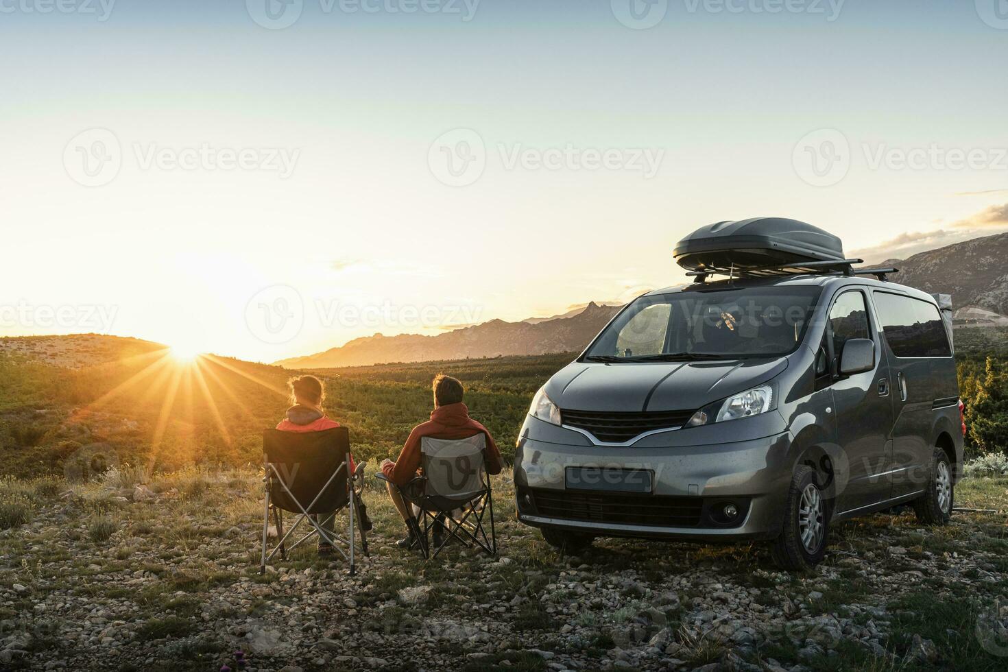 Man and woman sitting near camper van, looking at landscape at sunset photo