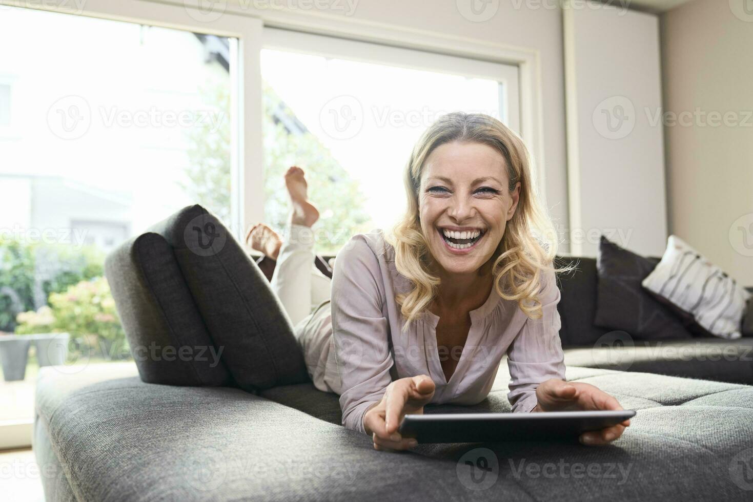 Portrait of laughing woman lying on couch at home using tablet photo