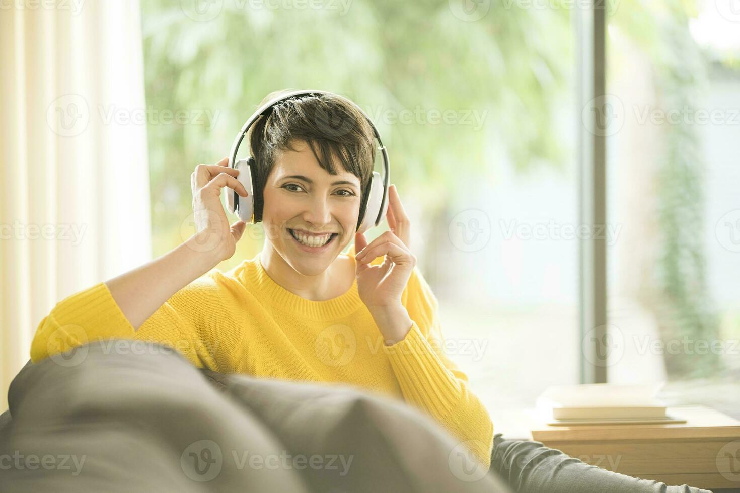 retrato de contento mujer sentado en el sofá a hogar escuchando música con auriculares foto