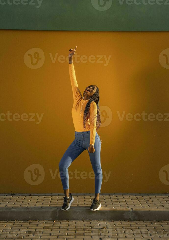 Teenage girl dancing with hand raised against yellow wall on street photo