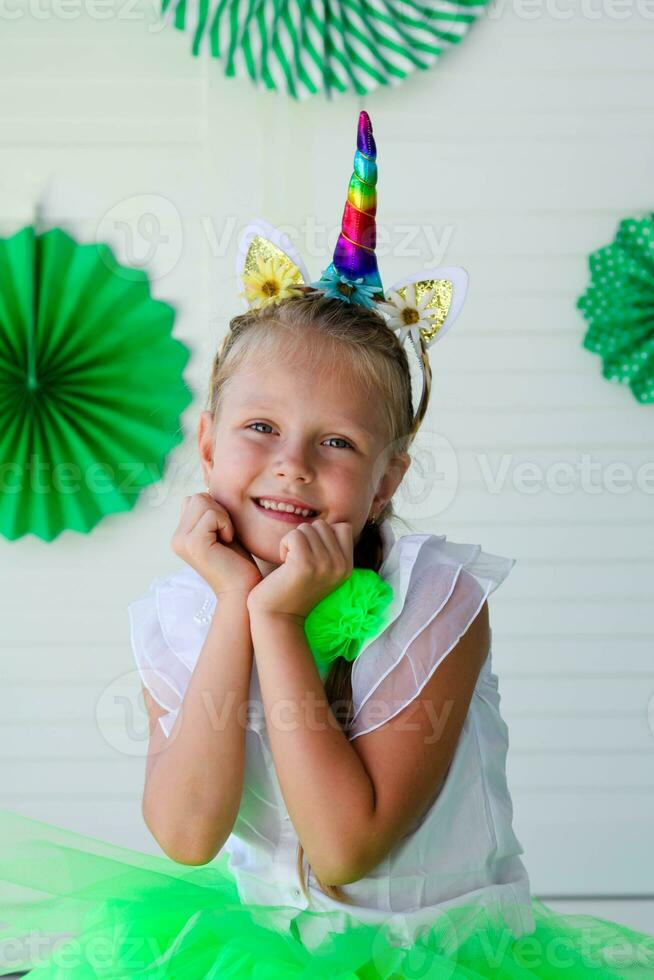 A little girl with a unicorn hoop on her head against a background of green photo decoration. Birthday for children.