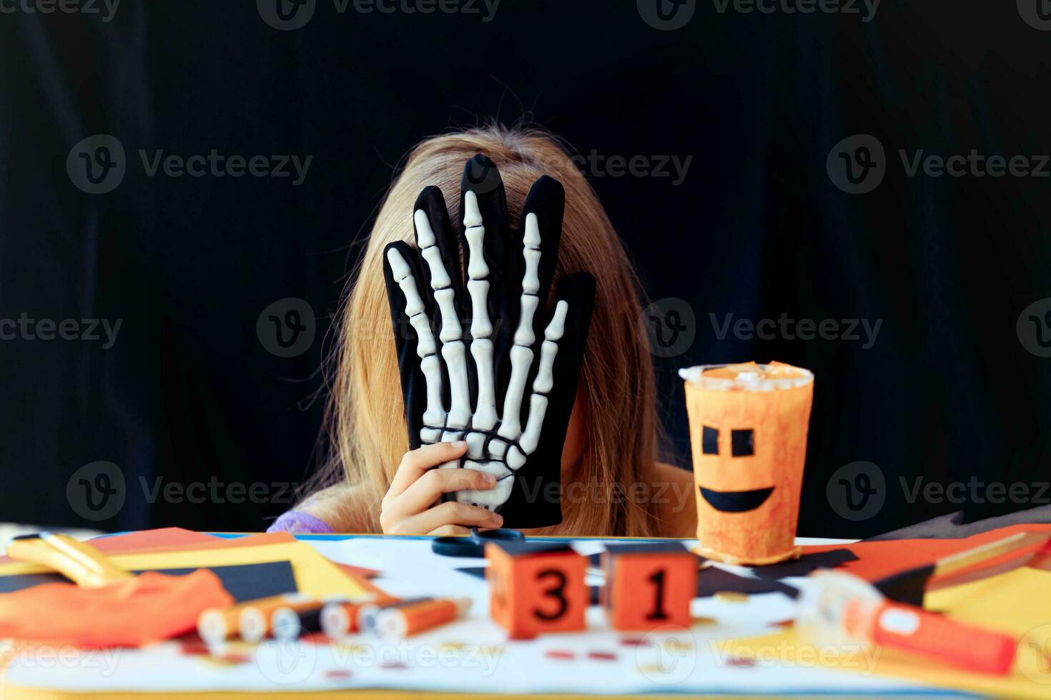 A little girl hides her head behind a skeleton glove on black background. Halloween holiday concept. photo