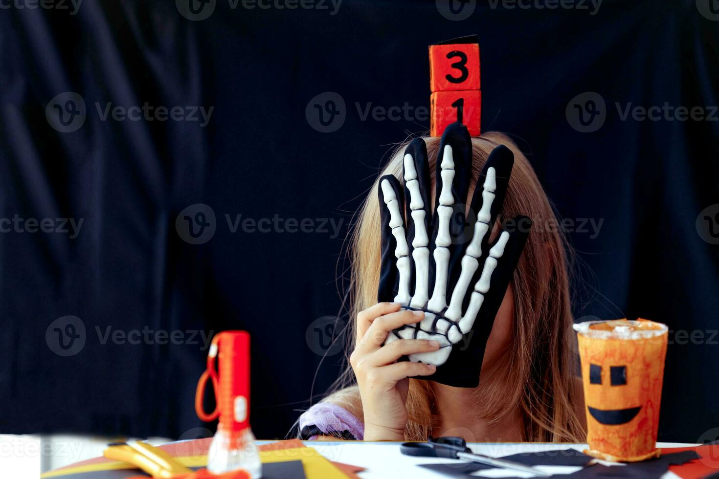 A little girl hides her head behind a skeleton glove, on her head is a wooden calendar with the date of October 31. photo