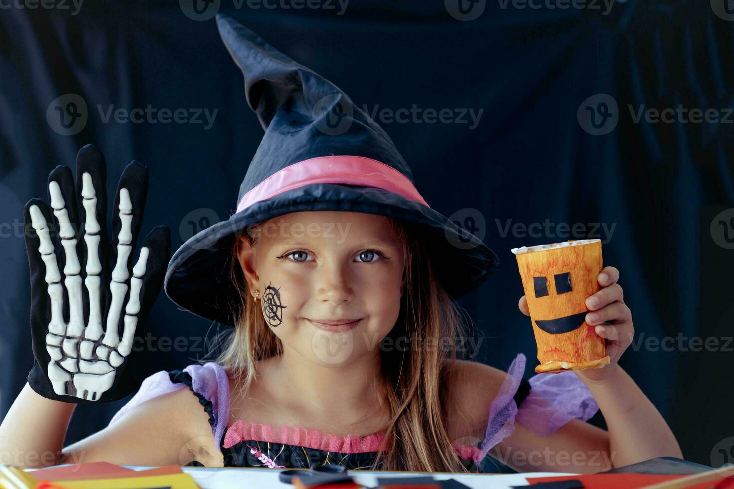 un pequeño niña en un bruja disfraz mira a el cámara y risas, participación un vaso calabaza y un esqueleto guante en su manos en negro antecedentes. foto