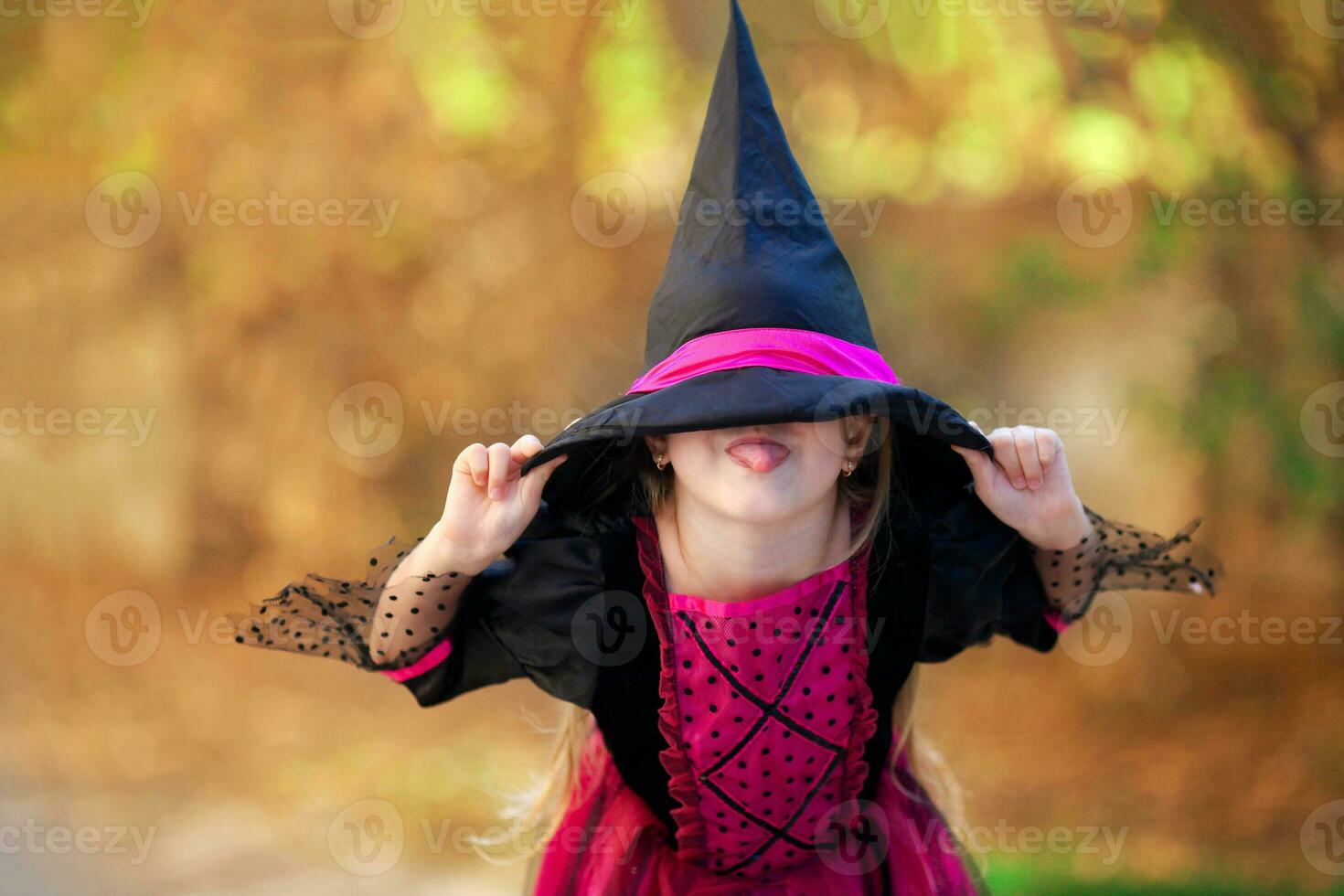 A portrait of a little girl in a witch costume hiding behind a black cap and showing her tongue. Halloween holiday. photo