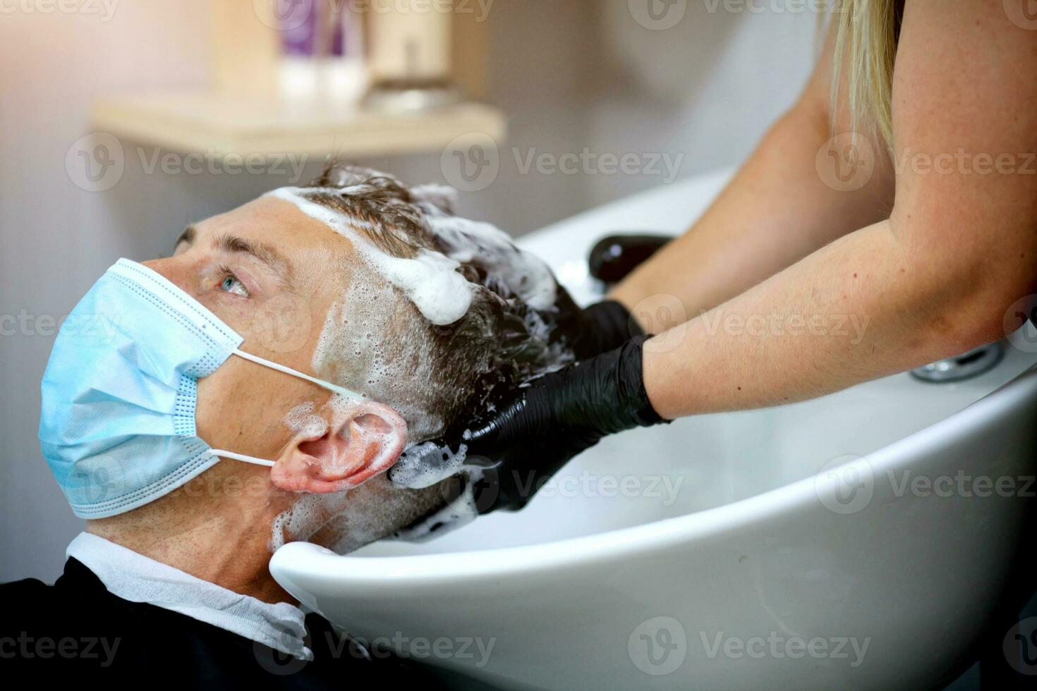 Female hairdresser hands in rubber gloves washes shampoo the head of a medical mask man. photo