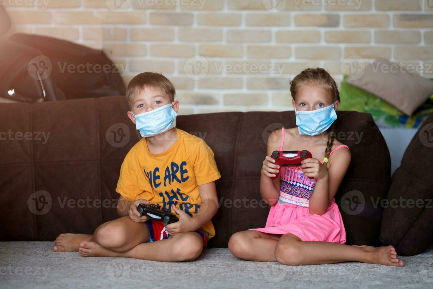 Little girl and boy play medical mask game console while sitting on sofa at home. photo