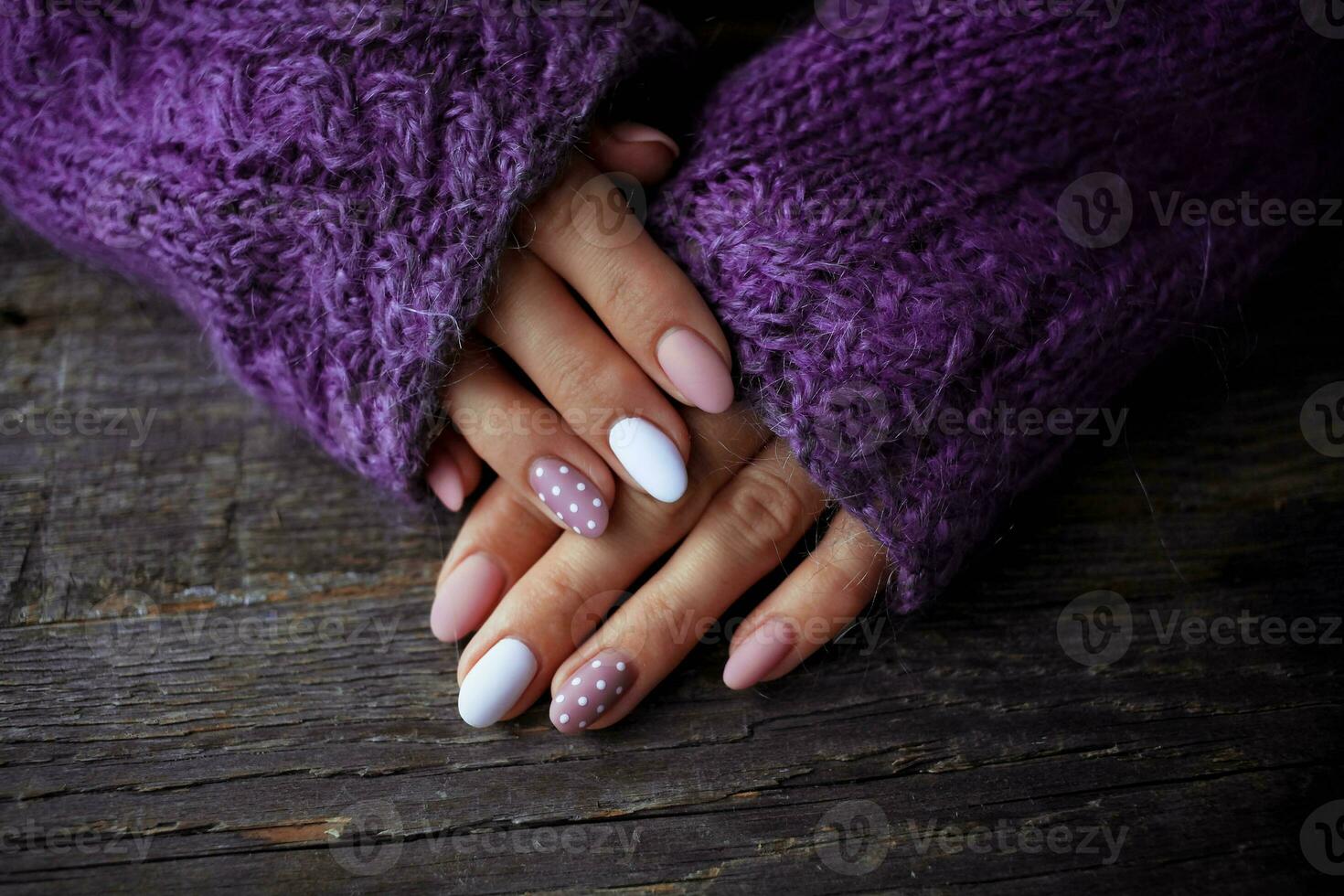 Women is hands with a beautiful manicure, in a violet knitted sweater on a wooden background in. Autumn trend, polish beige and white polka dots on nails with gel polish, shellac. photo