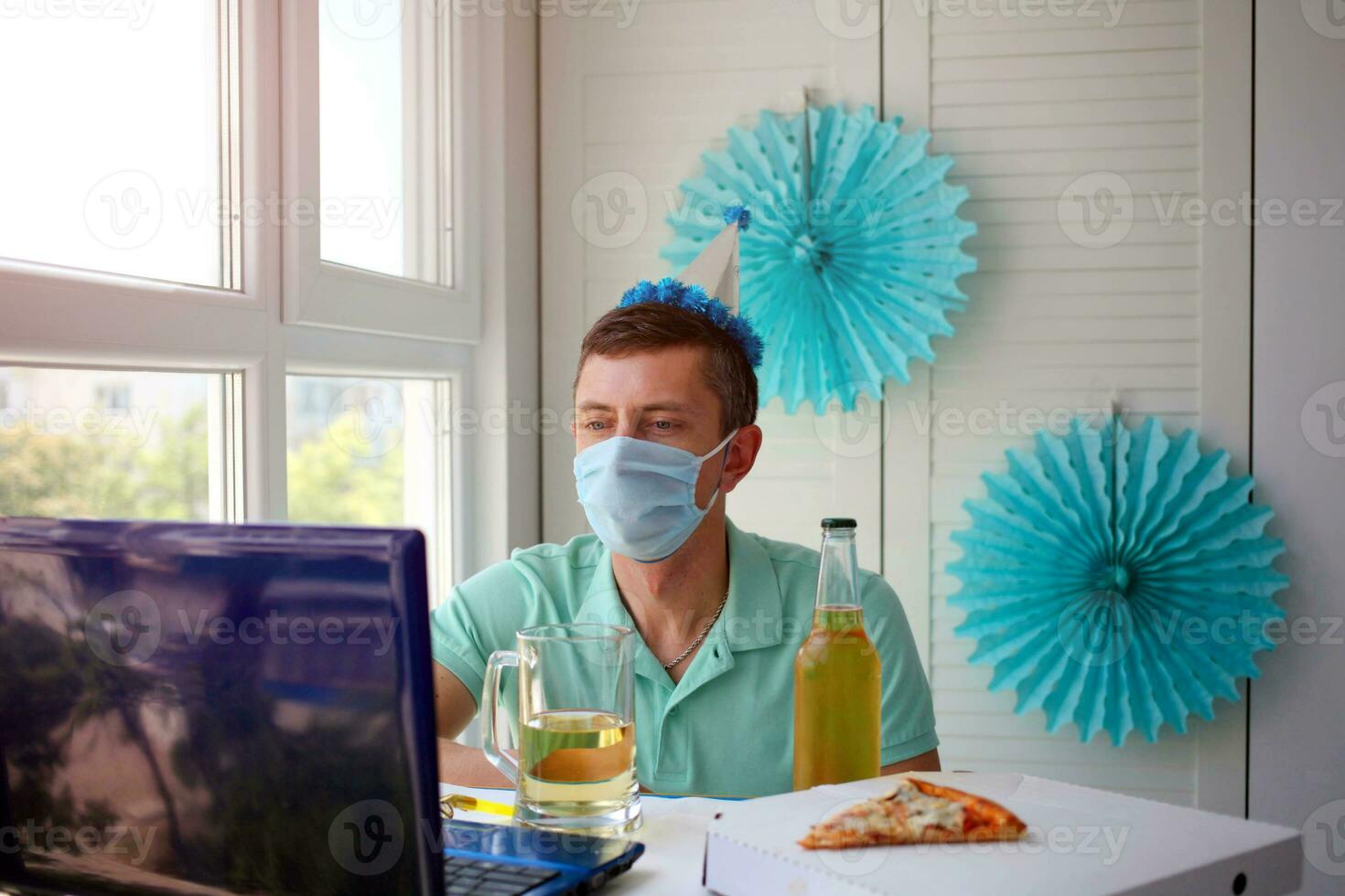 un hombre en un médico máscara se sienta en frente de un computadora portátil, celebra un cumpleaños con amigos terminado el Internet y sostiene un vaso de alcohol en su manos. foto