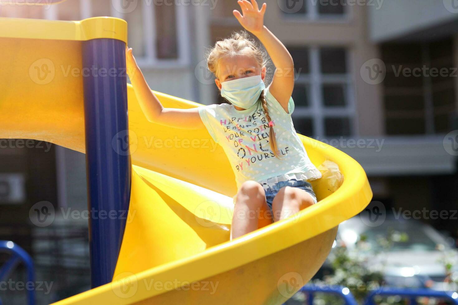A little girl in a medical mask is riding a slide  on the playground photo