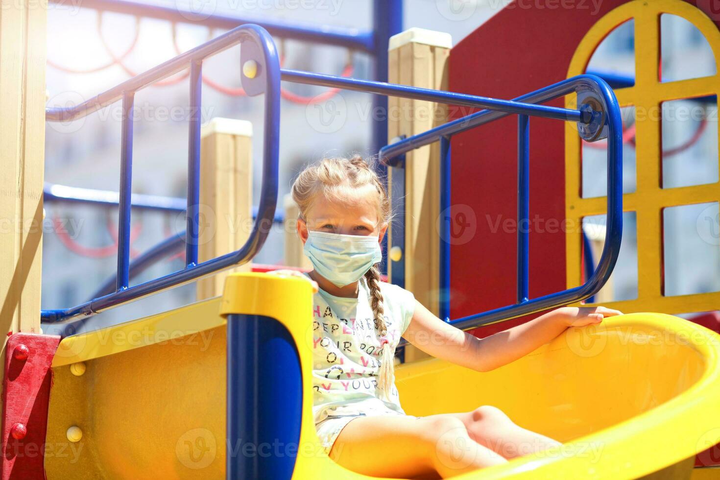 A little girl in a medical mask is riding a slide  on the playground photo