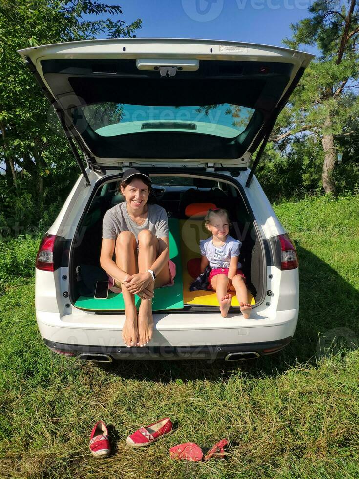 joven mamá con su pequeño hija sentado descalzo en un coche maletero en campo. contento familia yendo a gastar el noche en el blanco estación vagón en naturaleza. concepto de de viaje, descanso y la carretera viaje picnic foto