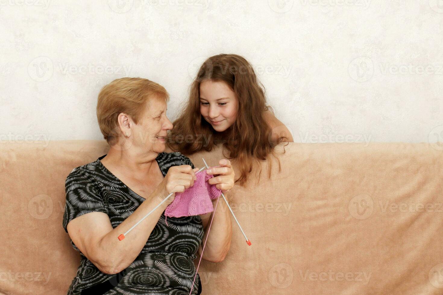 grandmother shows her granddaughter how she knits a warm pink sweater for her photo