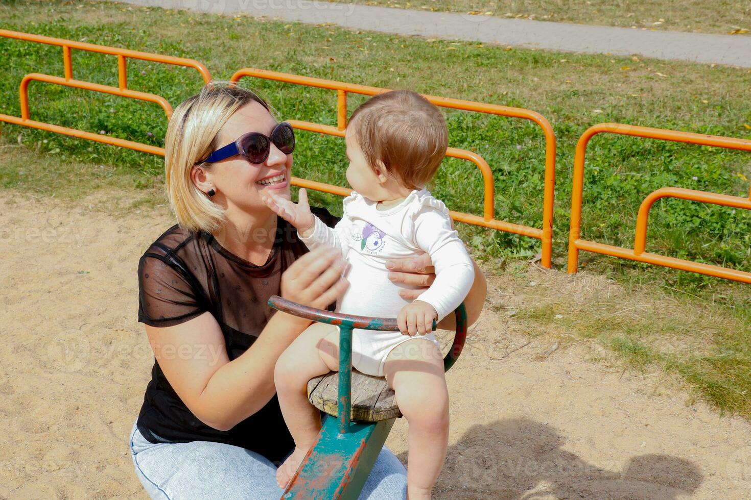 mom rolls baby daughter on a swing on the playground, talking with the child photo