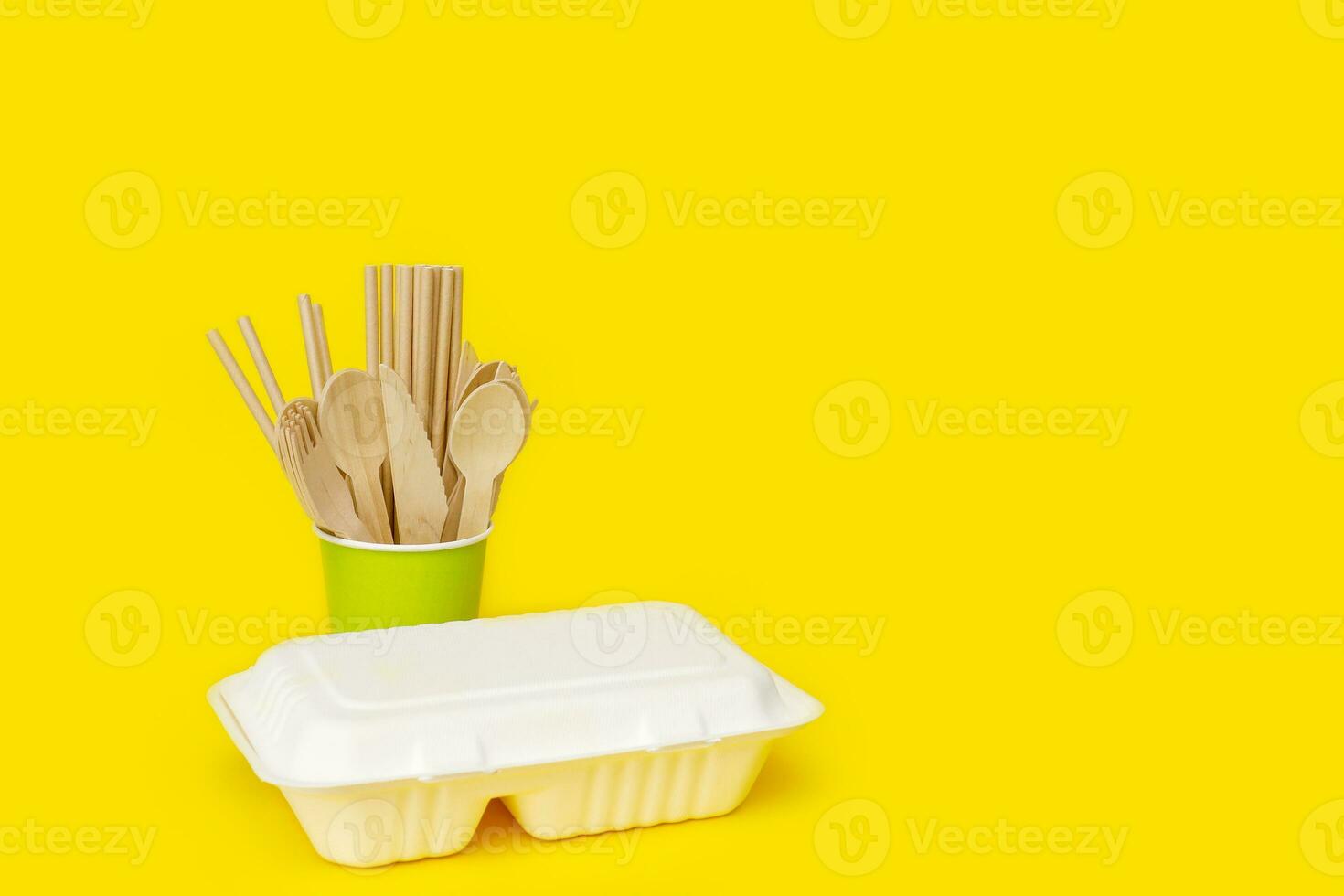 disposable wooden spoons, forks and knives in a paper cup with a corn raw lunchbox container on a yellow background with copy space, recycled photo