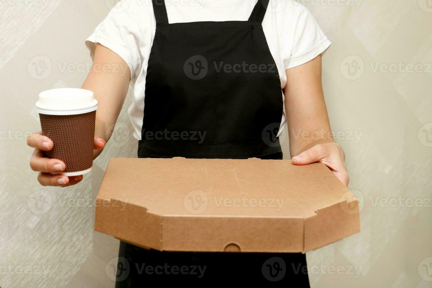 un mujer café trabajador sostiene fuera un caja con confeccionado Pizza y un taza de café foto