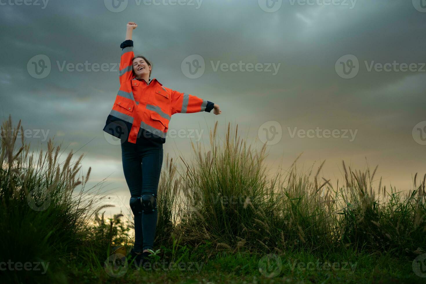 Young woman in engineer uniform and high visibility with raised arms standing on grassy field at sunset, The concept of relax time after work photo