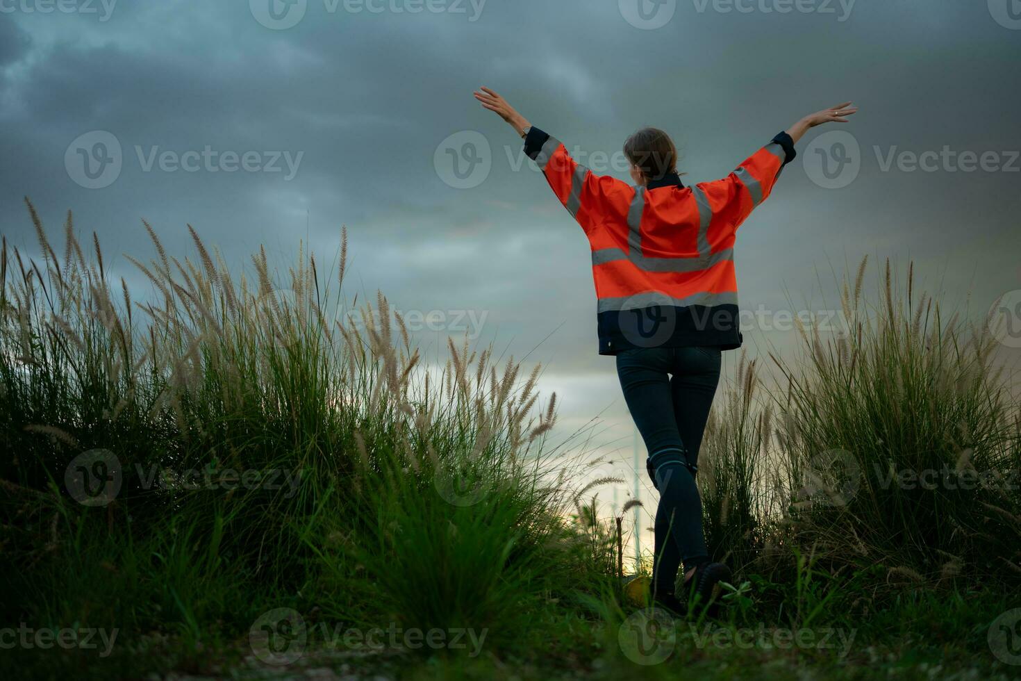 joven mujer en ingeniero uniforme y alto visibilidad con elevado brazos en pie en herboso campo a atardecer, el concepto de relajarse hora después trabajo foto