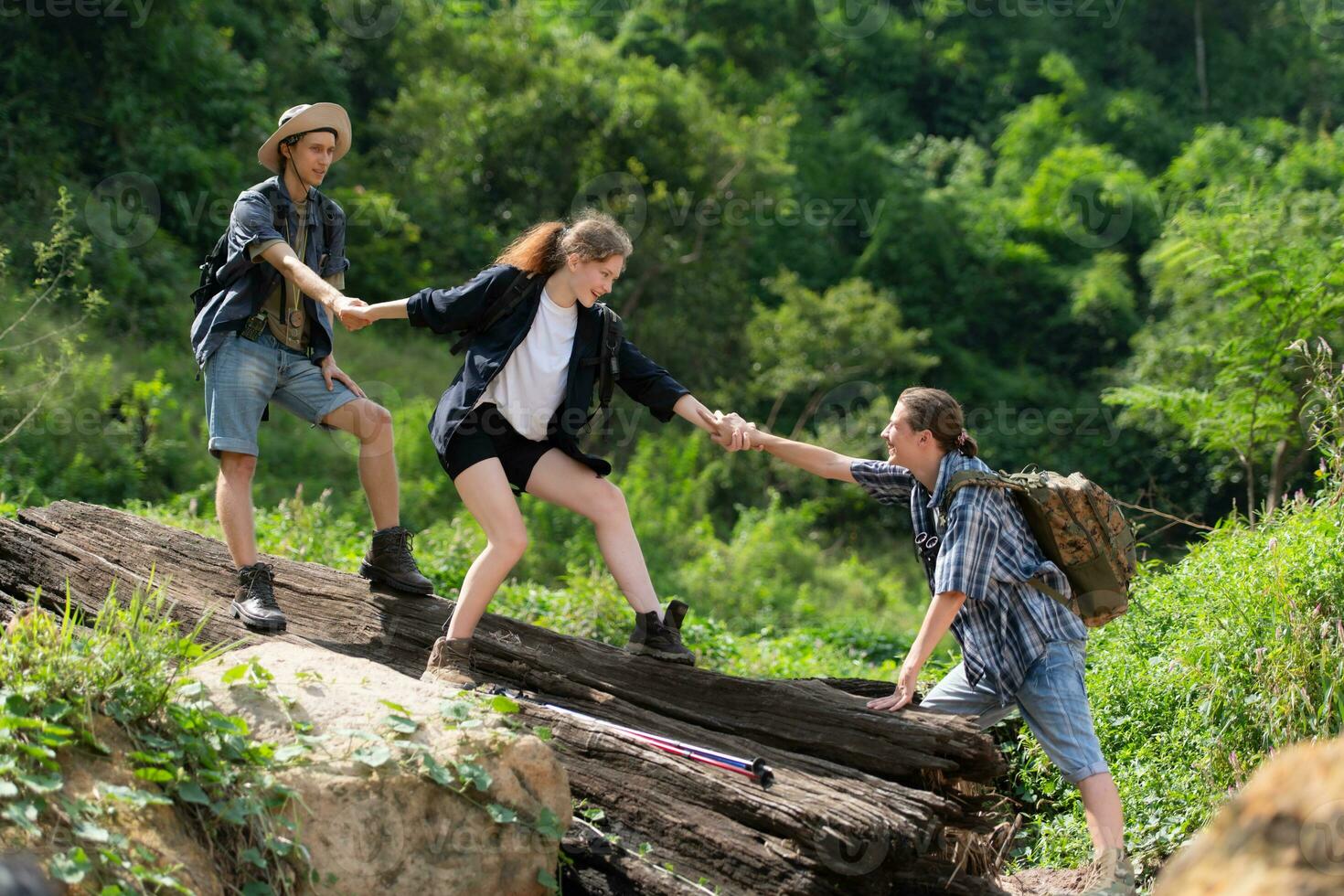 Group of friend on a sunny day in the forest Young group hiker with backpack and trekking poles helping to pull each other up to stand on the log photo