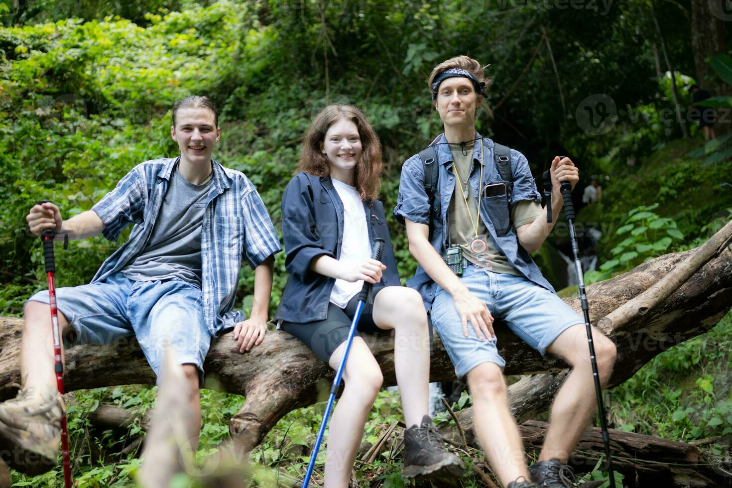 Group of friends with backpacks and sticks sitting on a fallen tree, Take a break during the hike photo