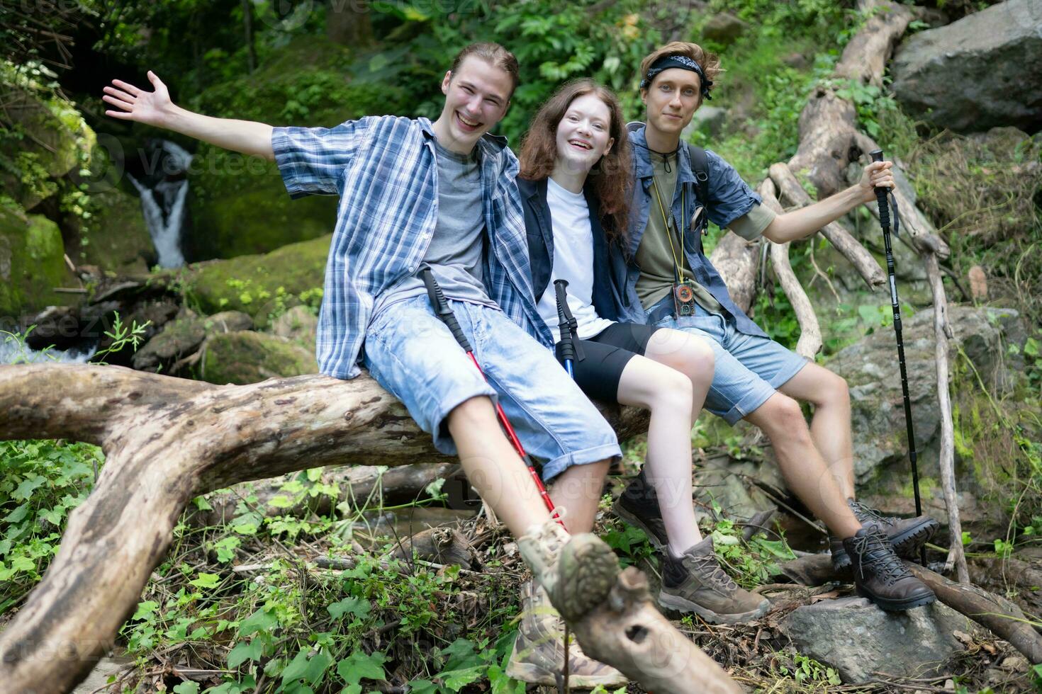 Group of friends with backpacks and sticks sitting on a fallen tree, Take a break during the hike photo